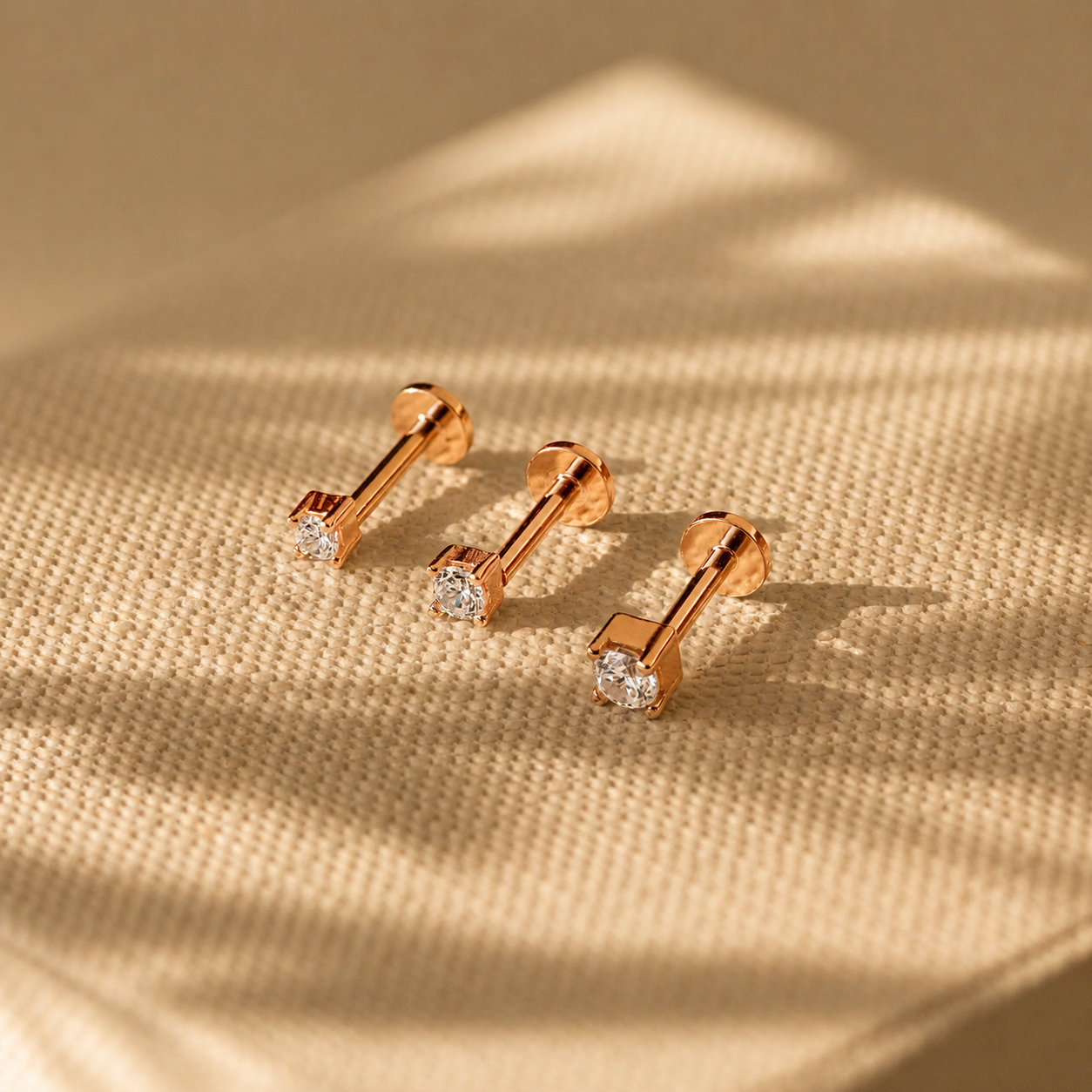 Rose gold barbell-shaped earrings with clear gemstones on a beige fabric background